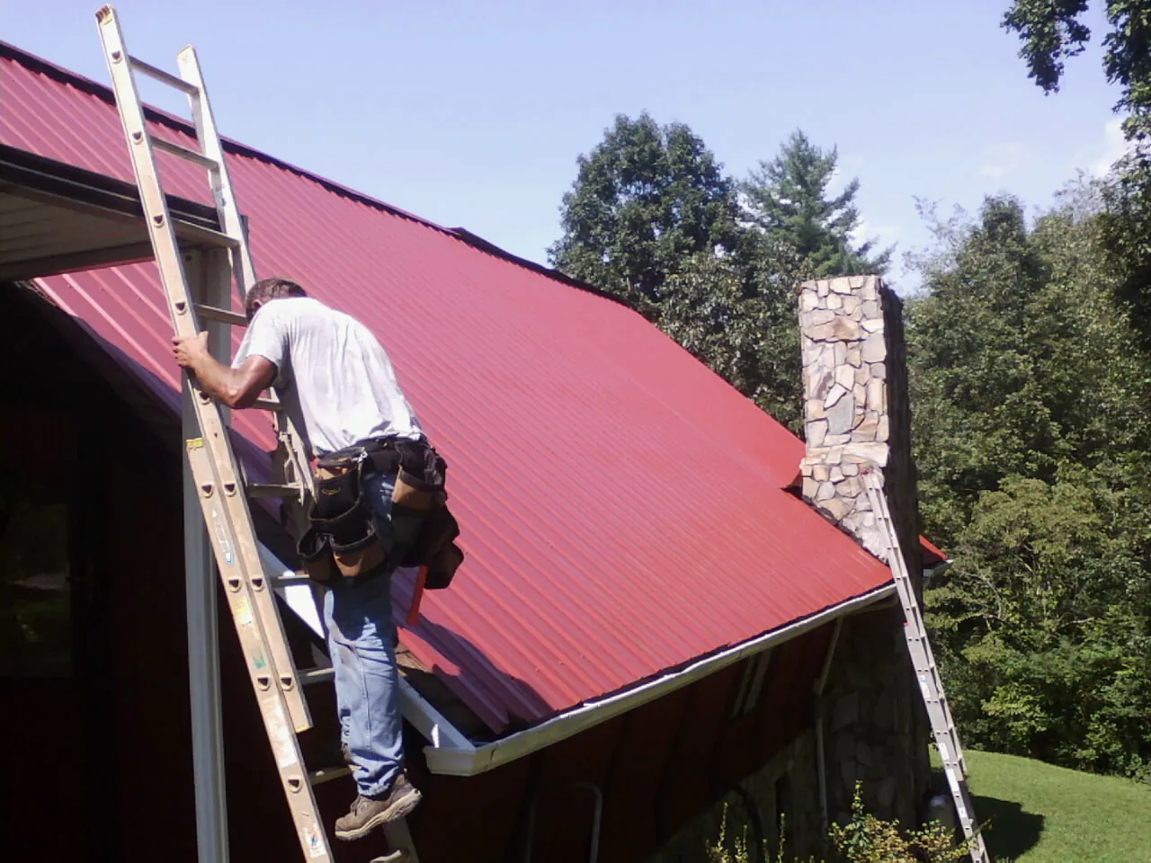 Roofer on a ladder beside a red metal roof and stone chimney — Soffit & Fascia Repair by Roofing Brothers in Garnet Valley